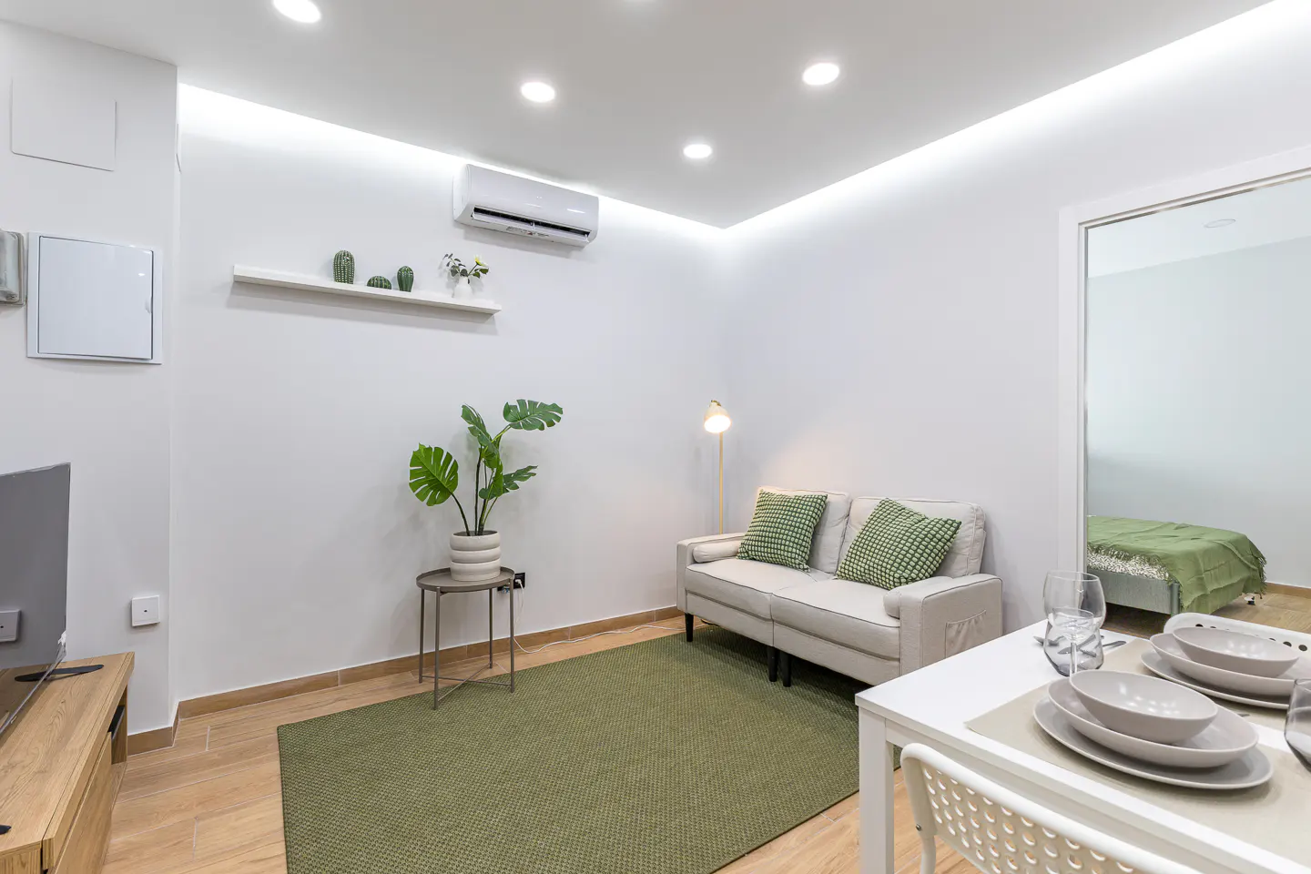 Bright living room with white walls, a beige sofa with green pillows, and a green rug. A dining table is set for two.