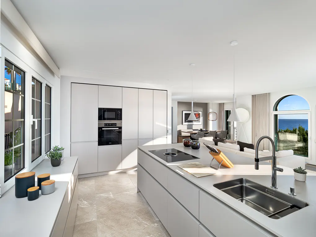 Bright, modern kitchen with white cabinets, island, and stainless steel sink. Ocean view through arched window. Open floor plan to living area.
