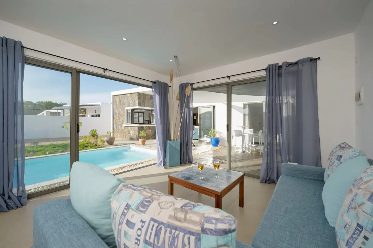 Bright living room with blue sofa, pillows, and curtains. Sliding glass doors open to a pool and patio with outdoor seating. A coffee table holds two wine glasses.