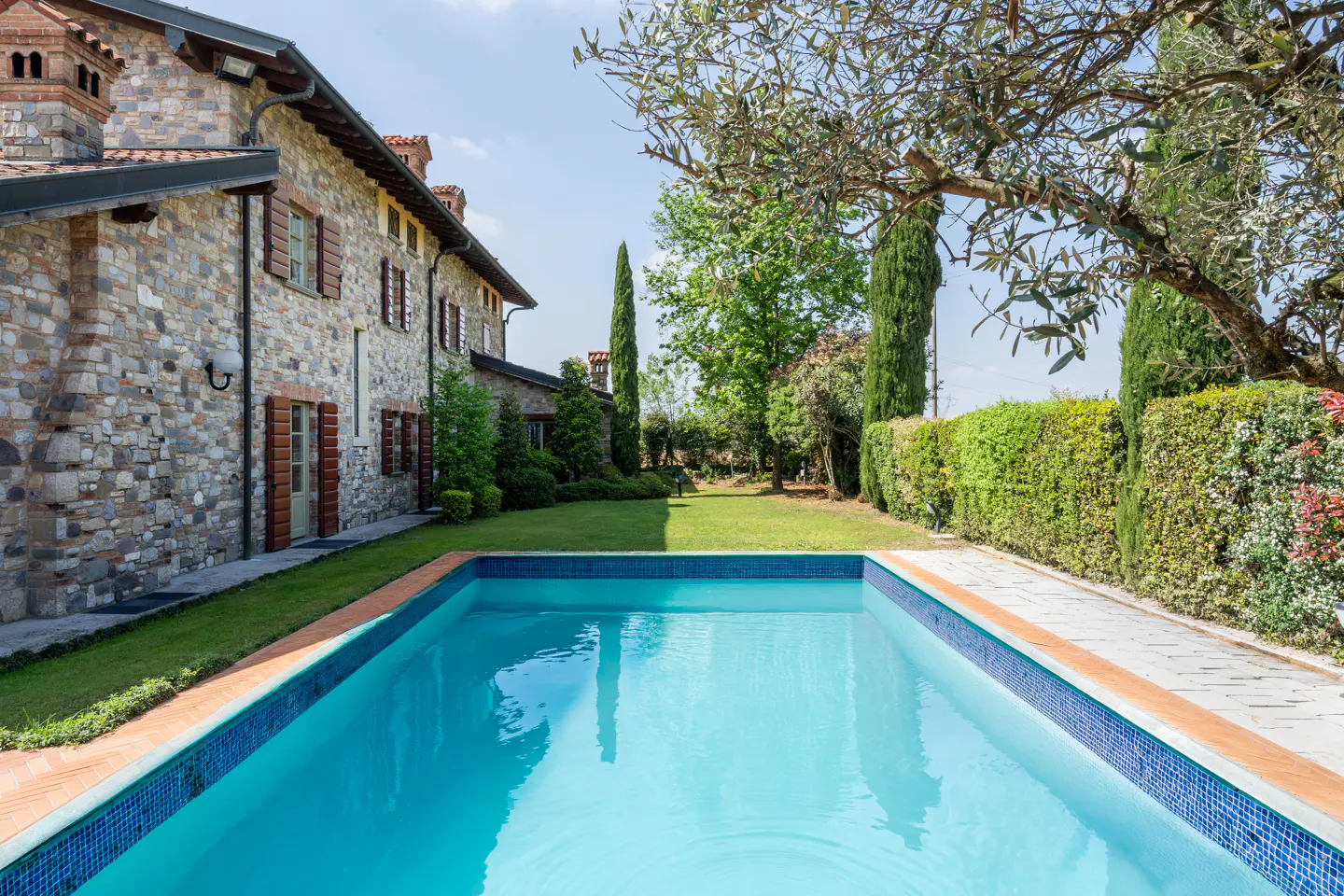 Exterior view of a stone house with a blue tiled pool, green lawn, and trees under a blue sky.