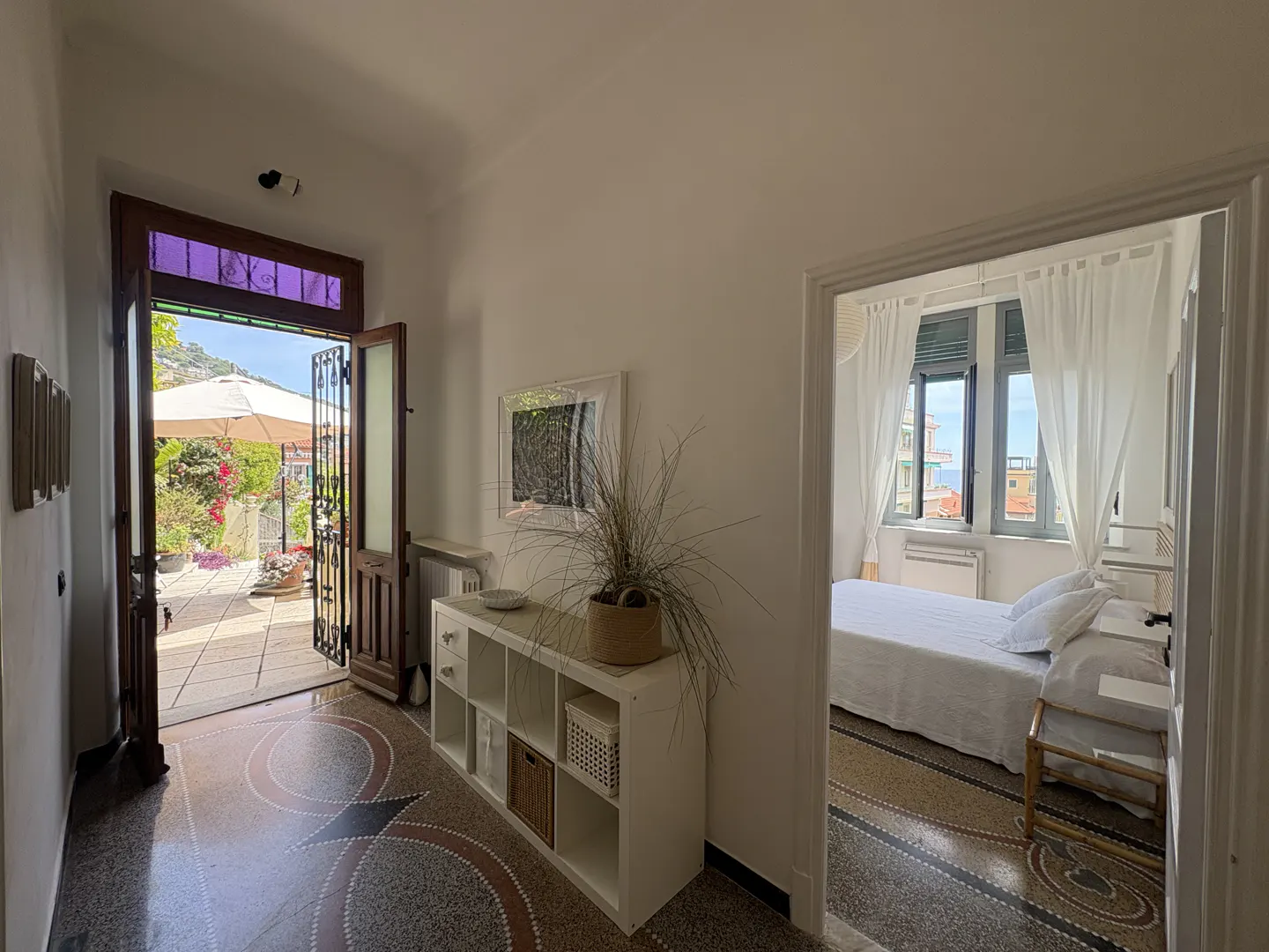 Hallway view to patio and bedroom. Open door to patio with umbrella, flowers. Bedroom with white bedding, sheer curtains, and ocean view.