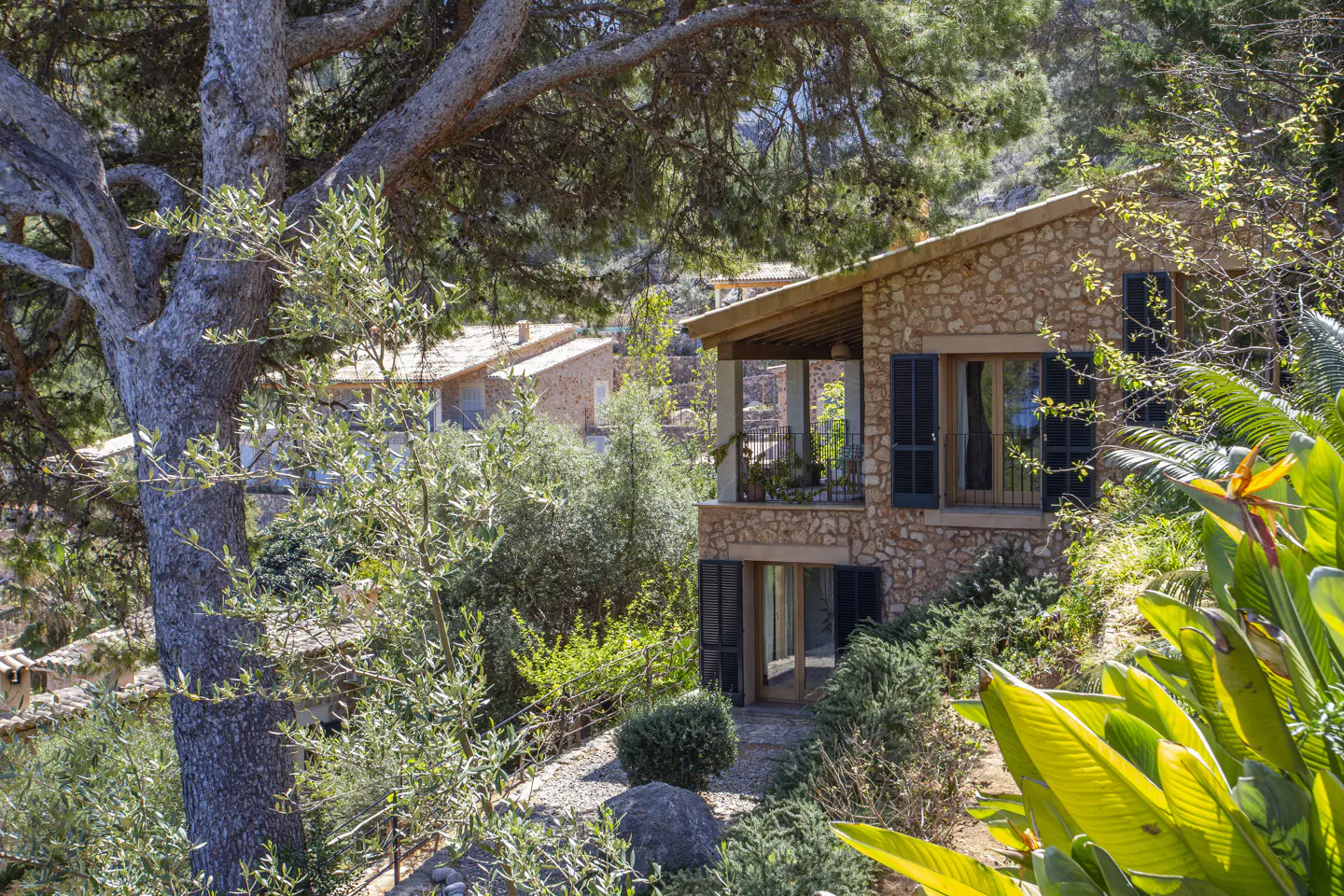 Stone house with dark shutters, surrounded by lush greenery and trees. A balcony with plants is visible.