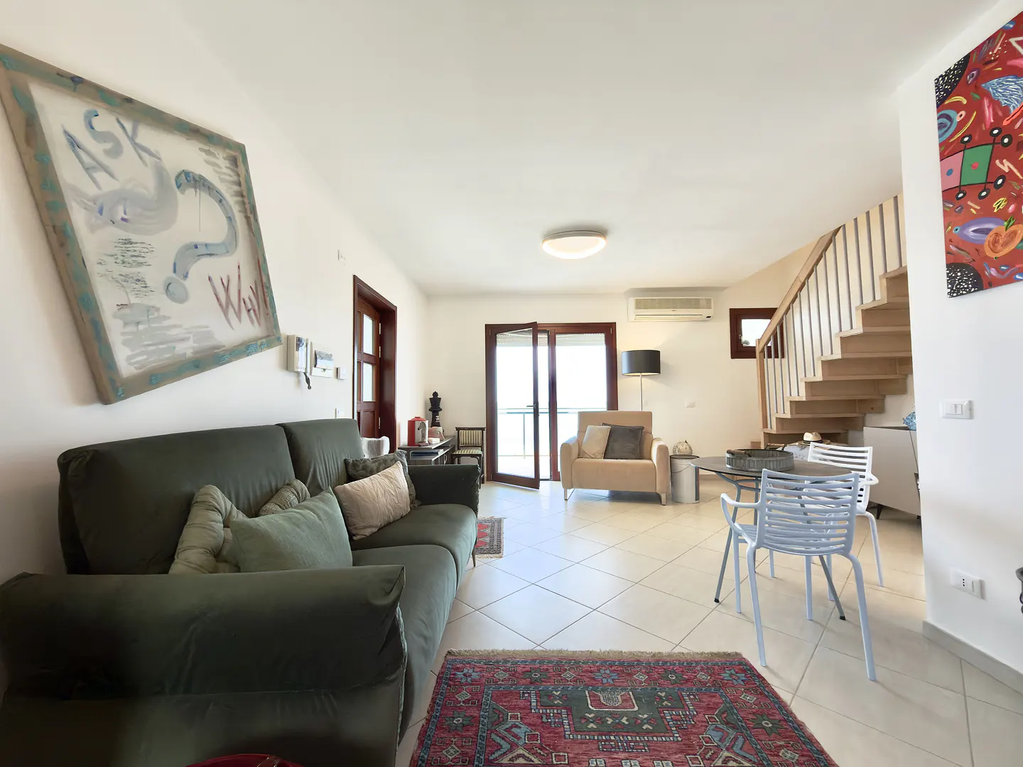 Bright living room with a green sofa, art, and stairs. A red rug sits on the white tile floor. Natural light streams in.