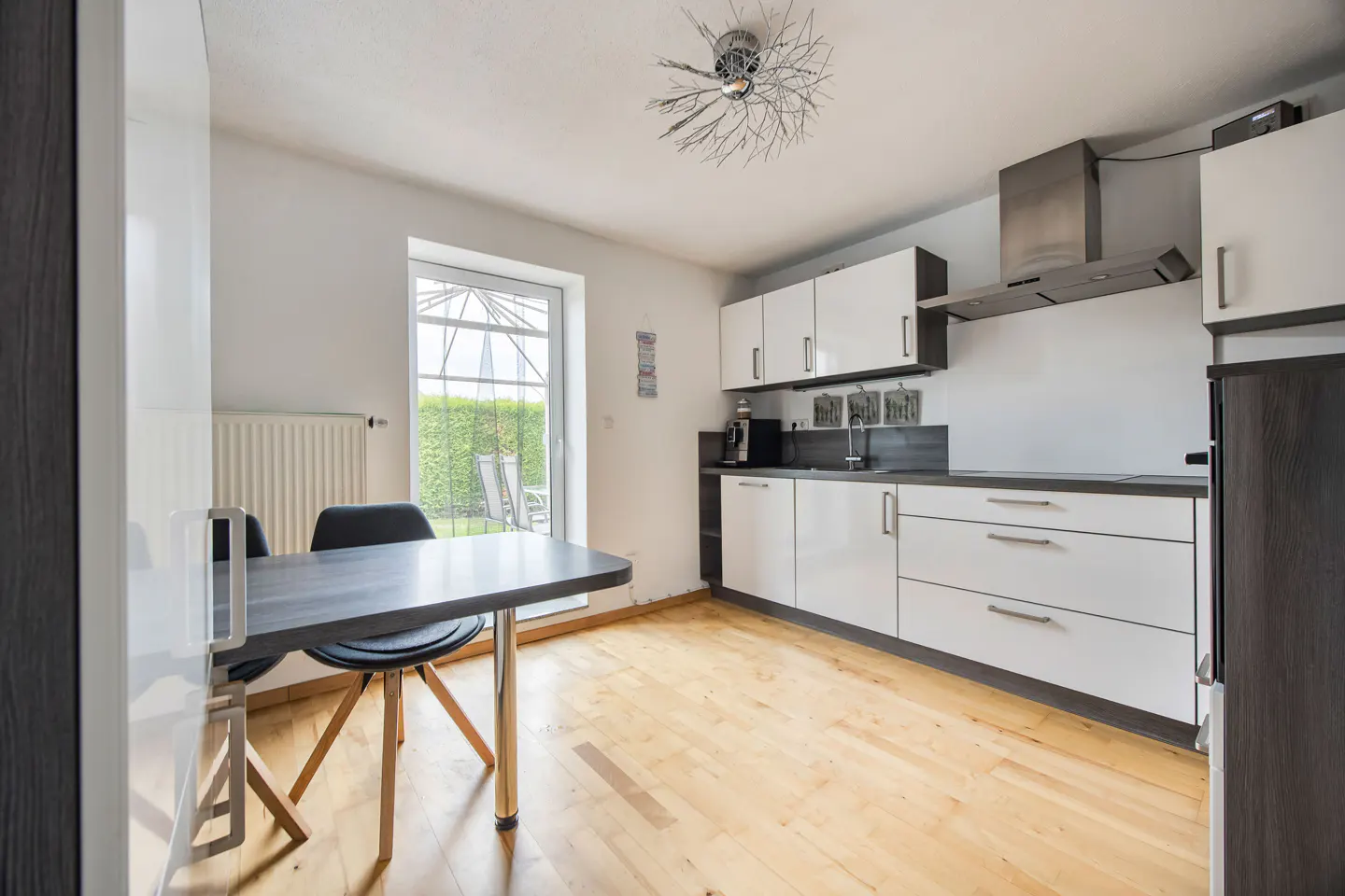 Bright kitchen with white cabinets, stainless steel appliances, and a gray table with black chairs. A door leads to a garden view.