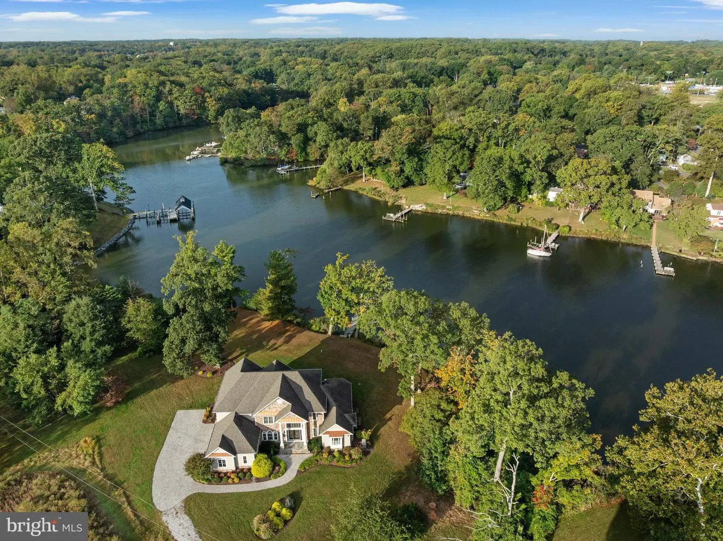 Aerial view of a tan and gray house with a dark gray roof on a green lawn near a river surrounded by green trees.