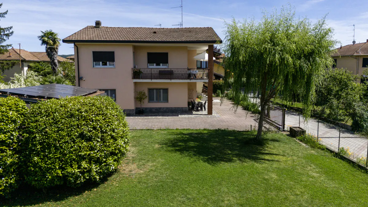 Two-story peach house with a brown roof, balcony, green lawn, and a large willow tree on a sunny day.