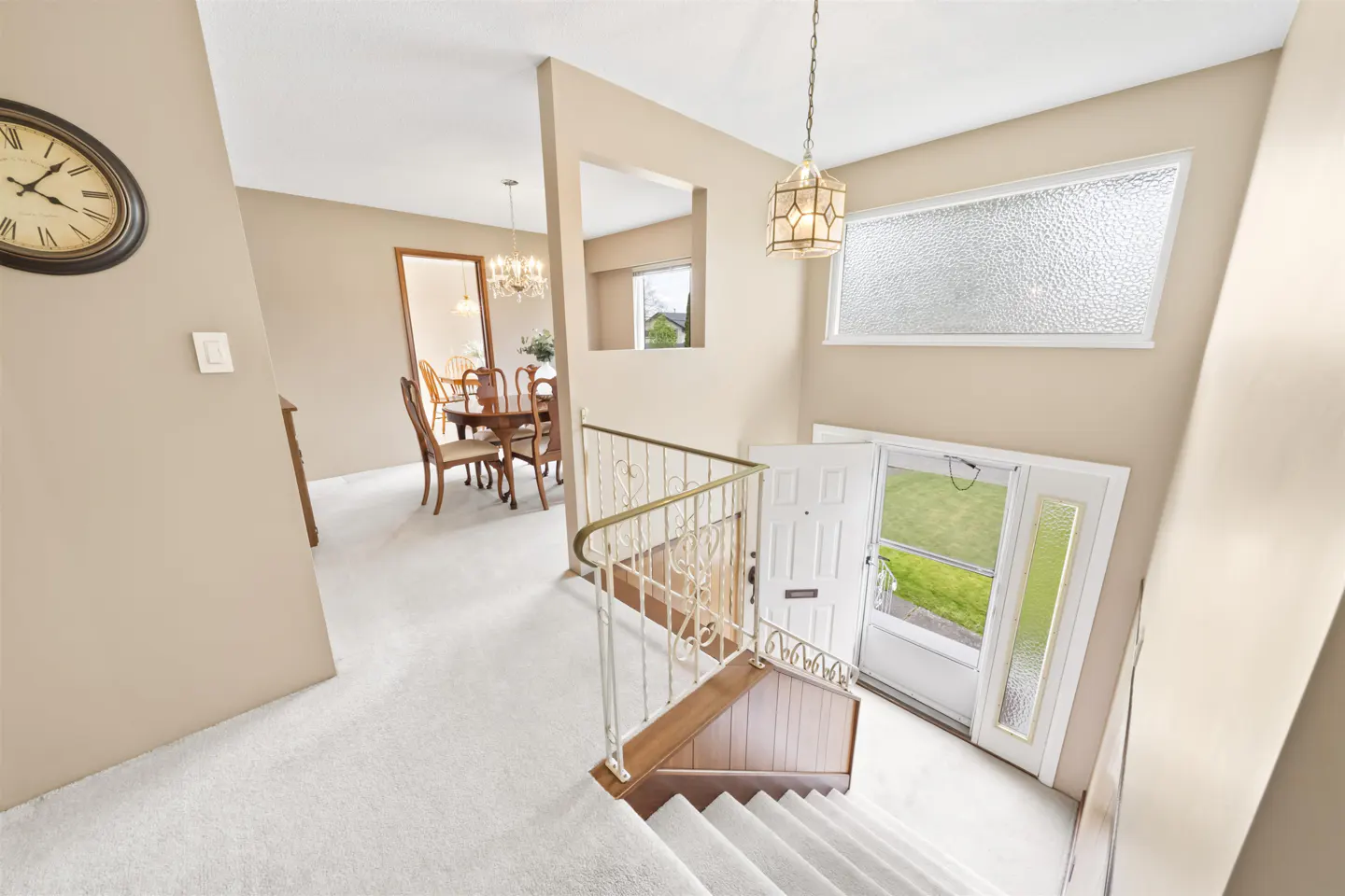 Foyer view from above shows stairs, white door open to green lawn, dining room, and beige walls. A clock hangs on the wall.