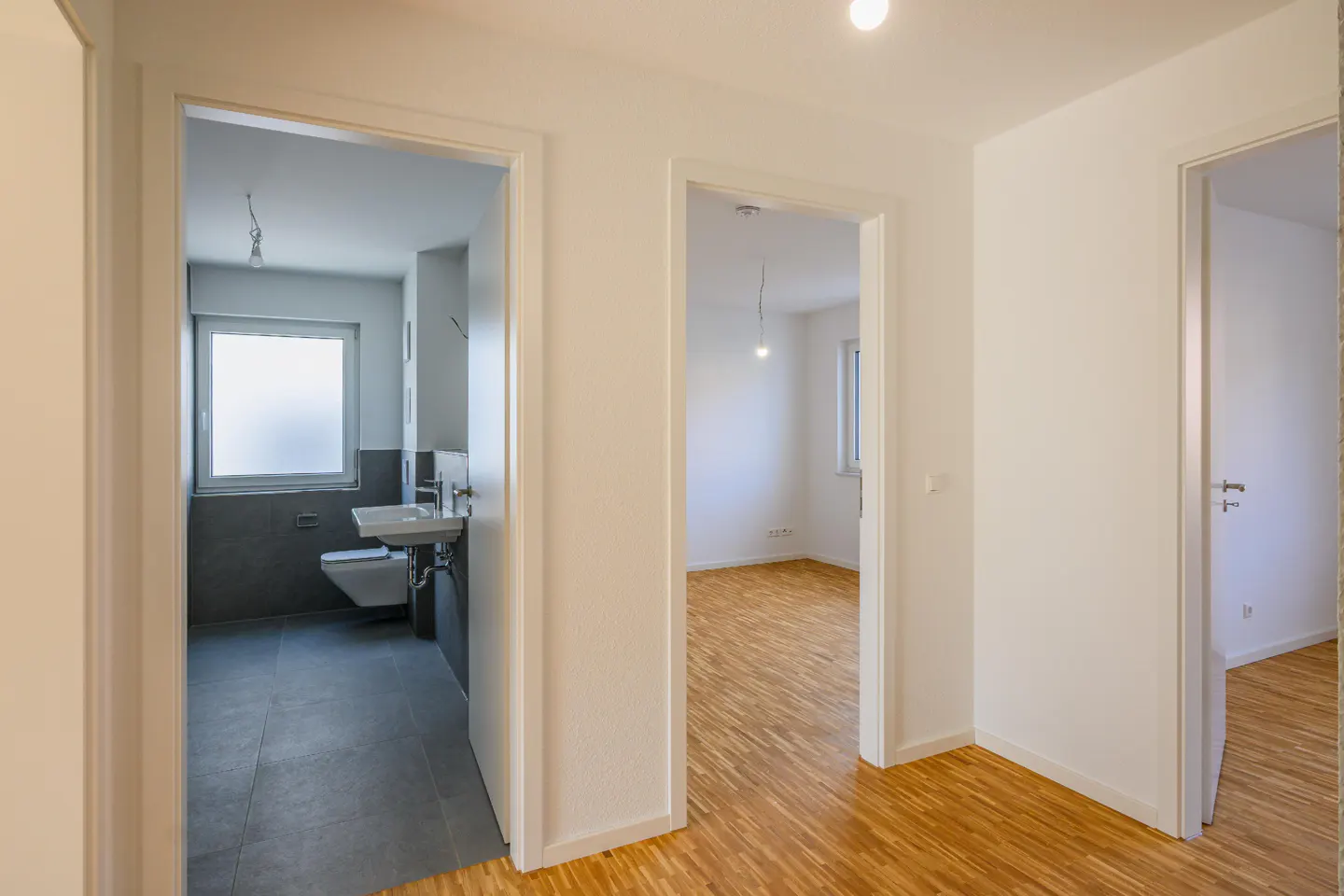 Hallway with white walls and wood floors. Open doorways lead to a gray bathroom and an empty room with a window.