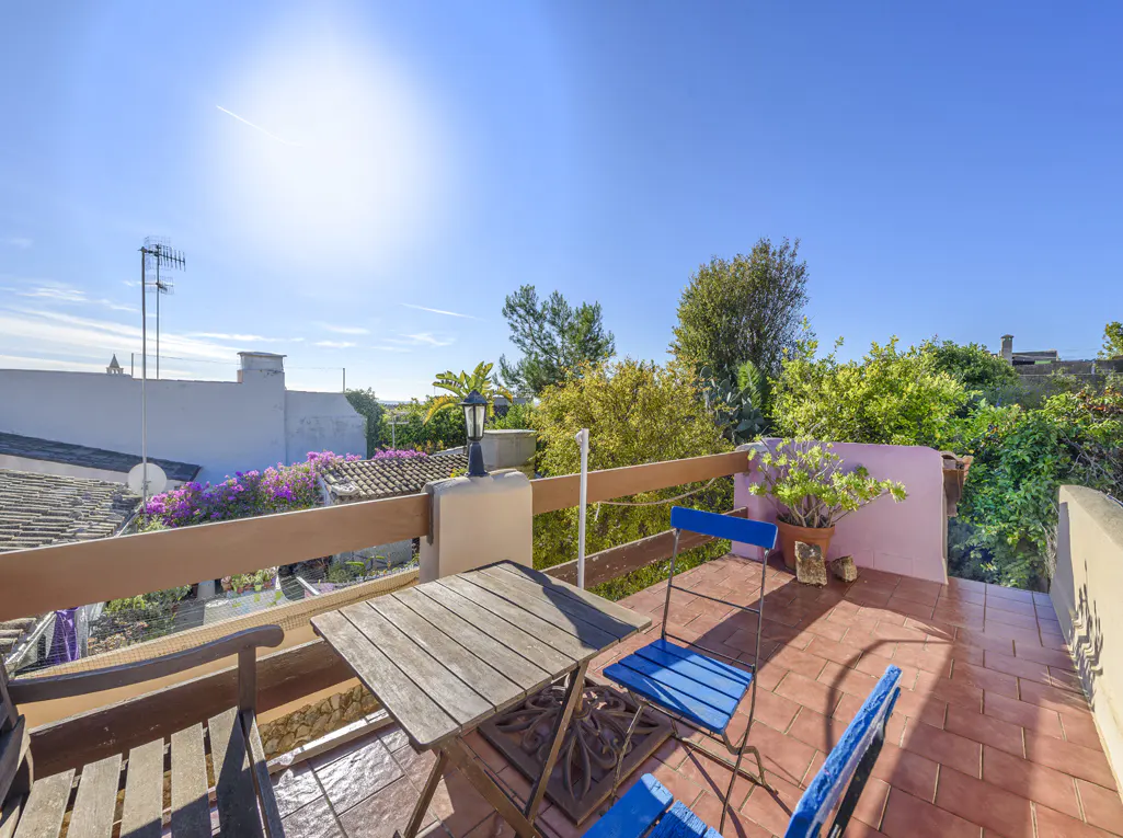 Outdoor patio with a wooden table and blue chairs on red tile. Lush greenery and a clear blue sky are in the background.