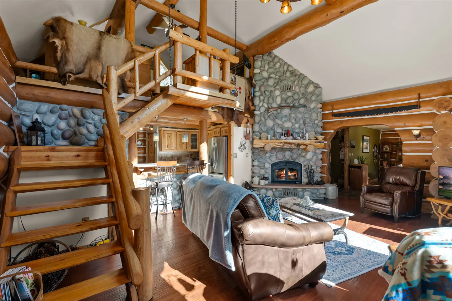 Rustic log cabin interior with stone fireplace, wooden staircase, and leather furniture. A mounted animal hide is visible above the stairs.