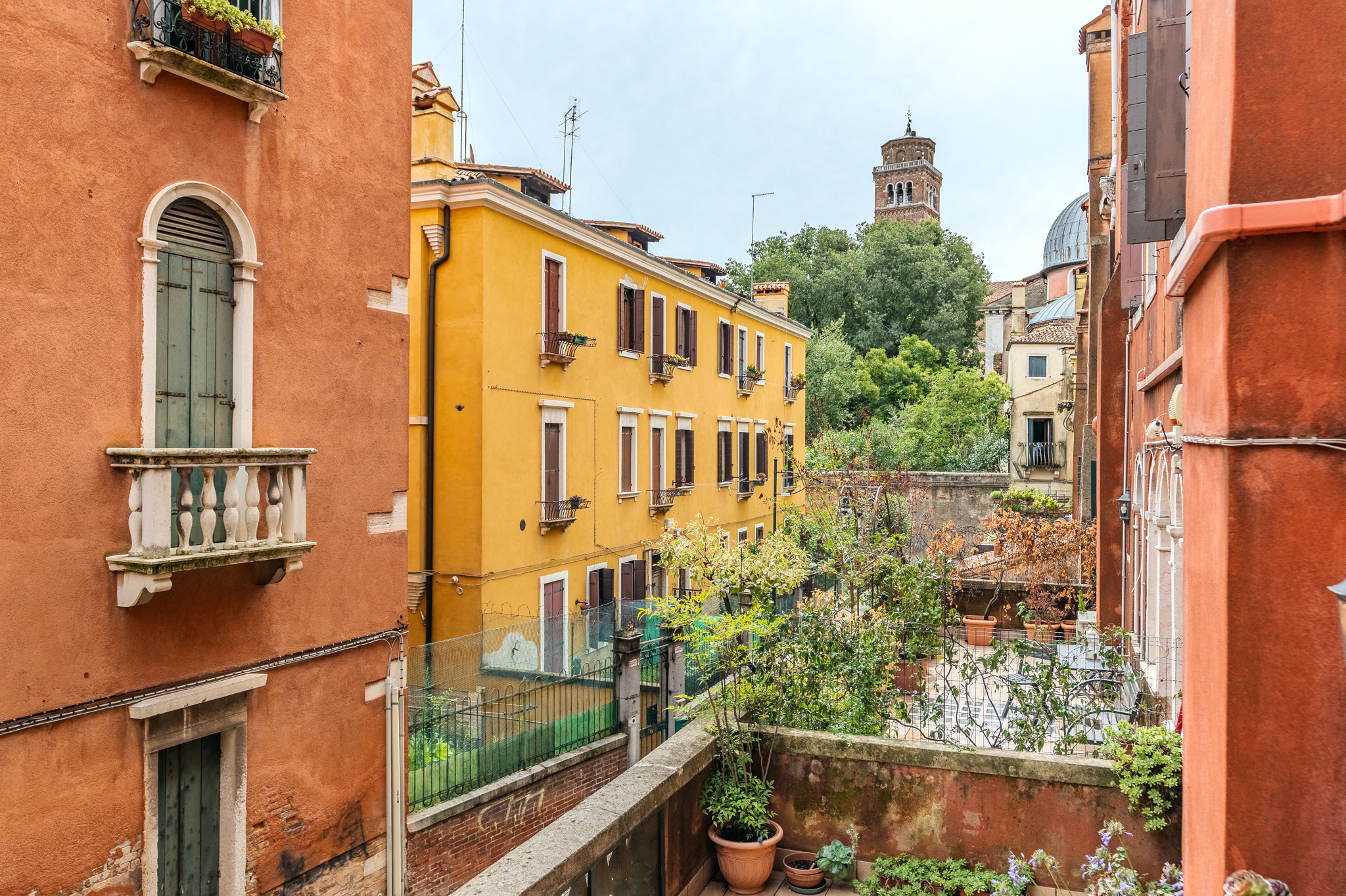 View of a yellow Venetian building and a garden from a balcony framed by terracotta-colored walls and a bell tower.