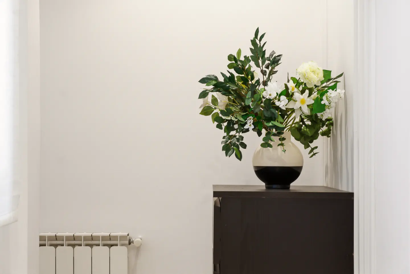 Interior shot of a room with a black cabinet holding a vase of white flowers and greenery against a white wall. A radiator is visible on the left.