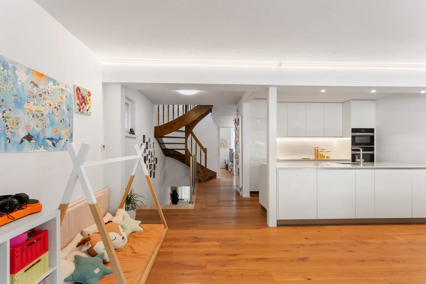 Bright, modern home interior with wood floors, white walls, and a wooden staircase. A child's teepee and kitchen are visible.