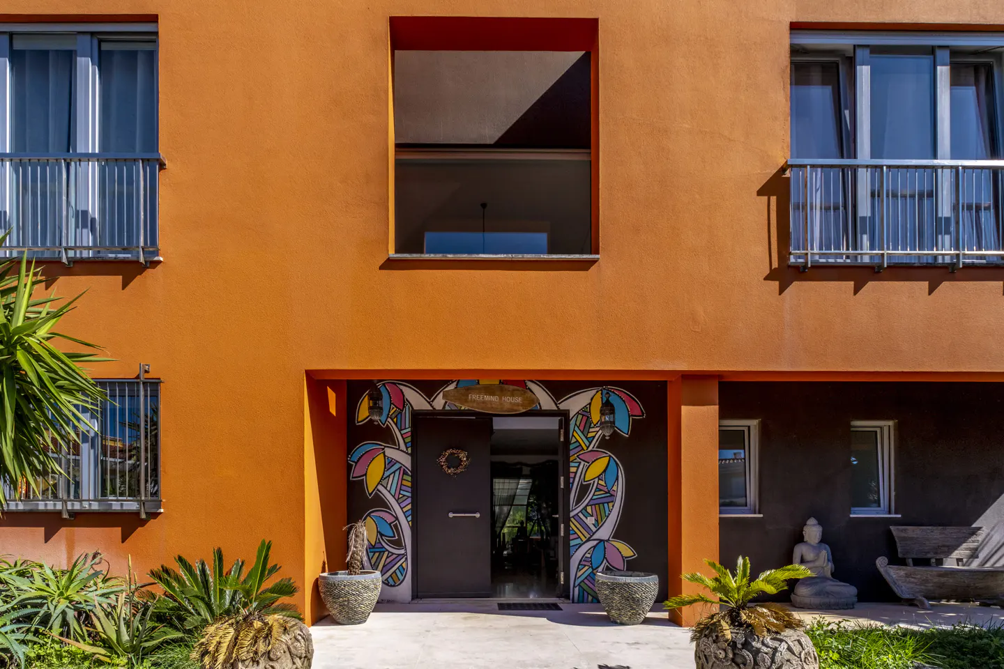 Orange building with open door, flanked by colorful floral murals. Windows above, plants and stone pots below.