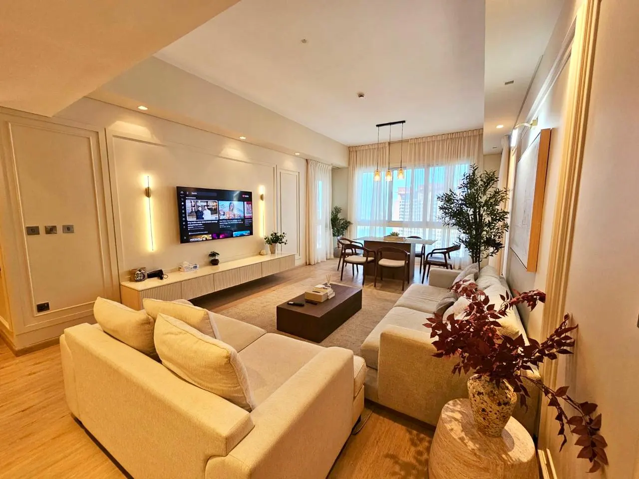Bright, modern living room with beige sectional sofa, wood floors, and a dining table set by a large window. A TV is mounted above a light wood console.