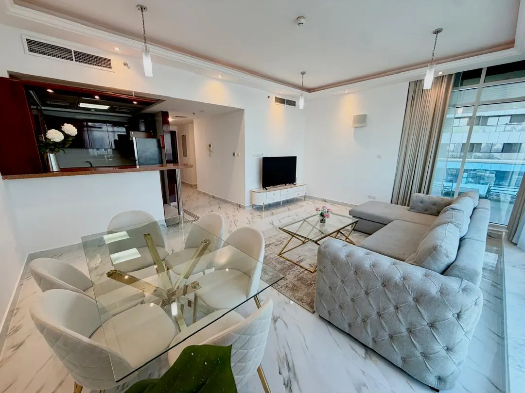 Modern living room with a glass dining table, tufted grey sectional sofa, marble floors, and an open kitchen area.