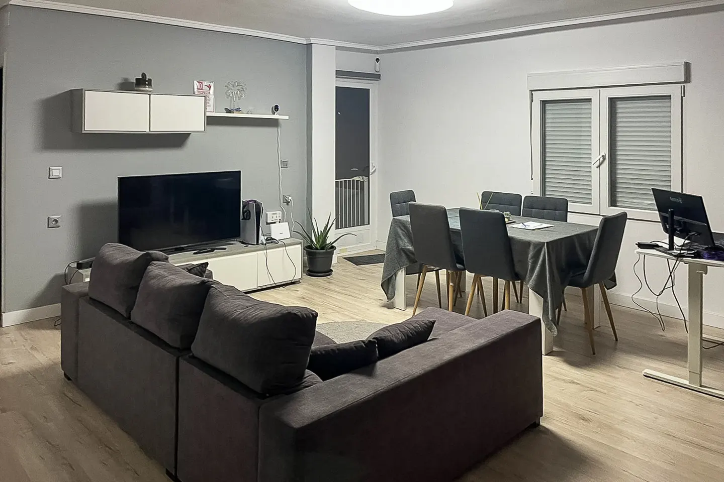 A modern living room with gray walls, a dark gray sofa, a dining table with six chairs, and a TV.