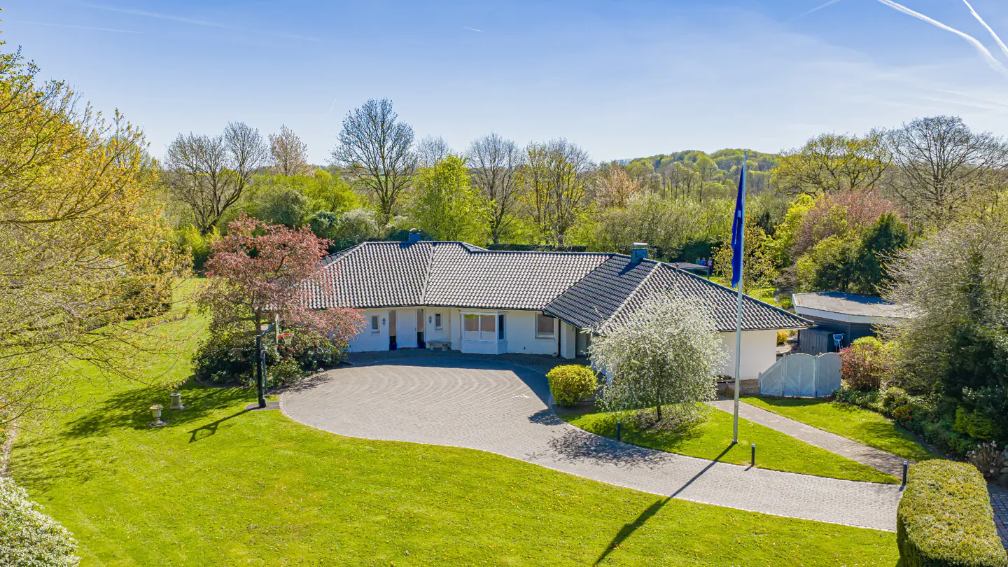 Aerial view of a white single-story house with a gray roof, surrounded by green lawn and trees under a blue sky.