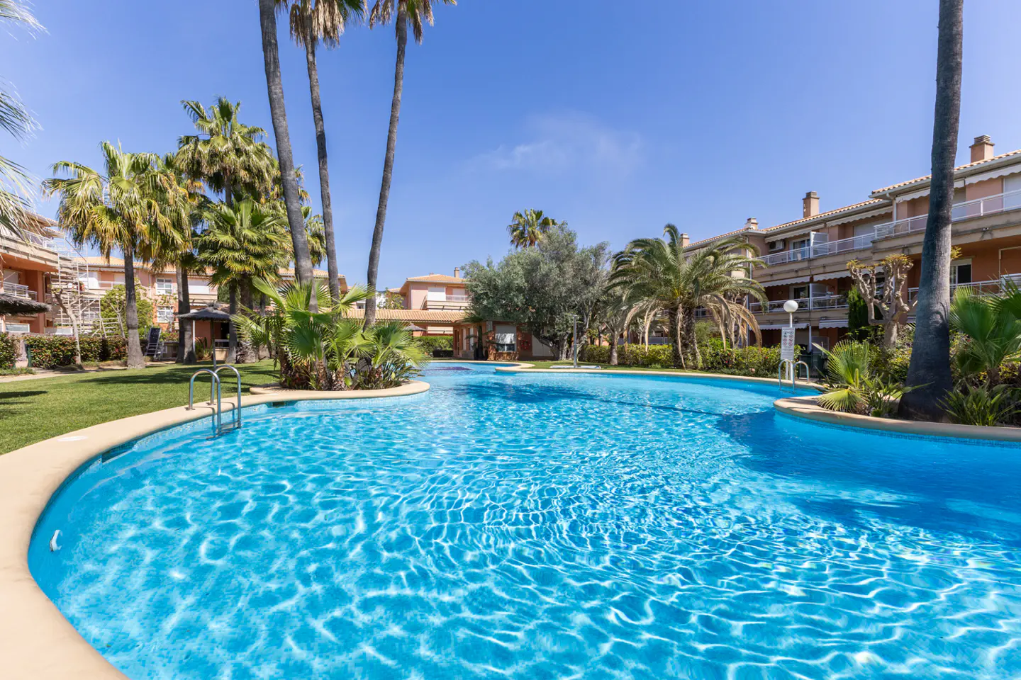 A turquoise pool surrounded by palm trees and buildings under a clear blue sky.