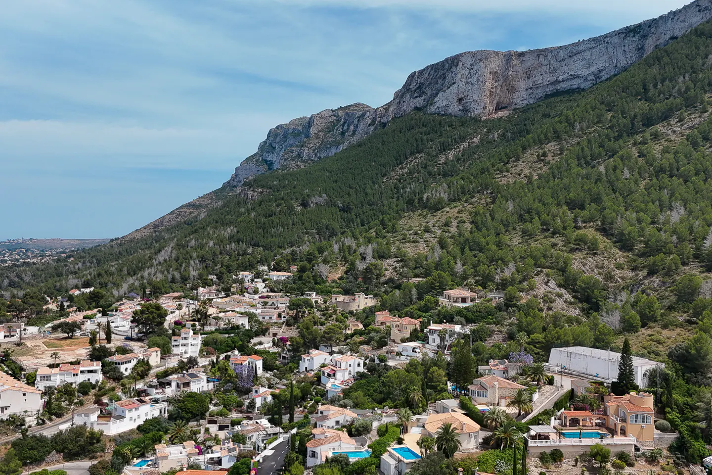 Aerial view of a Mediterranean hillside town with white villas, swimming pools, and lush trees beneath a rocky mountain.