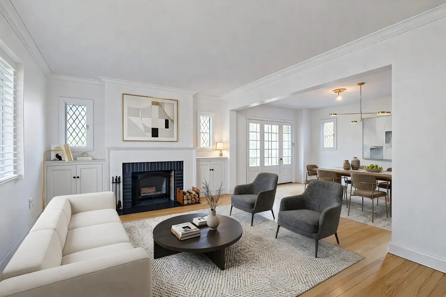 Bright living room with white walls, a fireplace, a cream sofa, two gray armchairs, and a round coffee table on a patterned rug. Dining room visible in the background.
