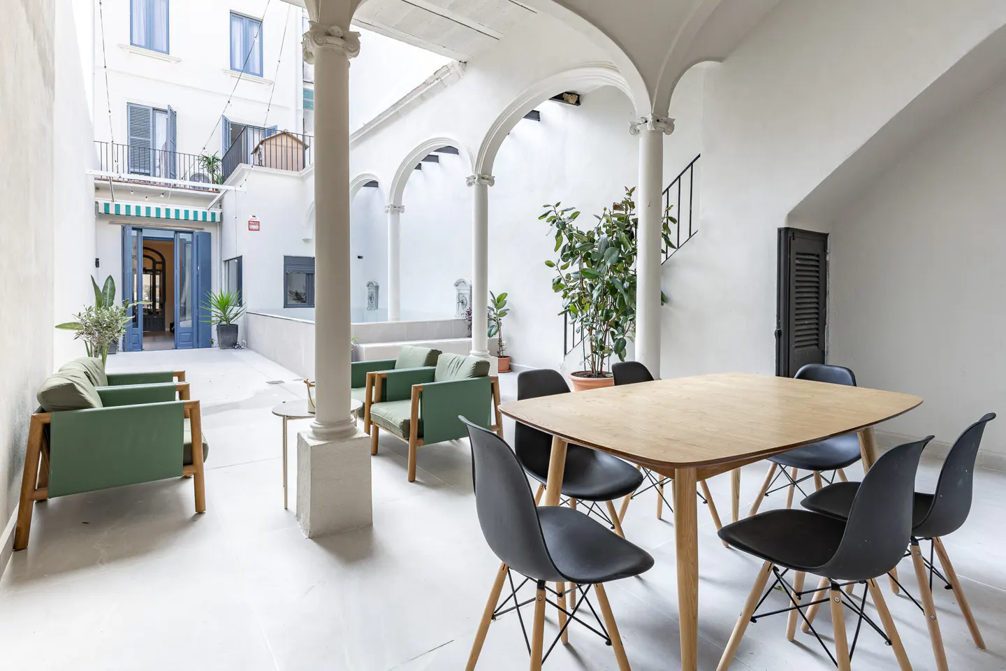Bright courtyard with green chairs, a light wood table with black chairs, and white walls with arched columns.