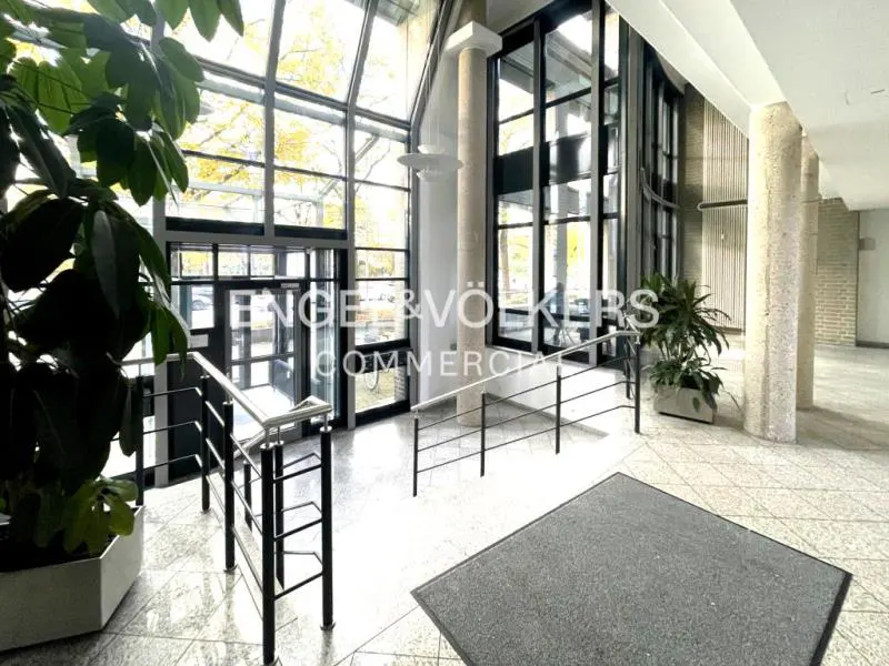 Interior view of a commercial building lobby with large windows, plants, and a staircase with black railings.