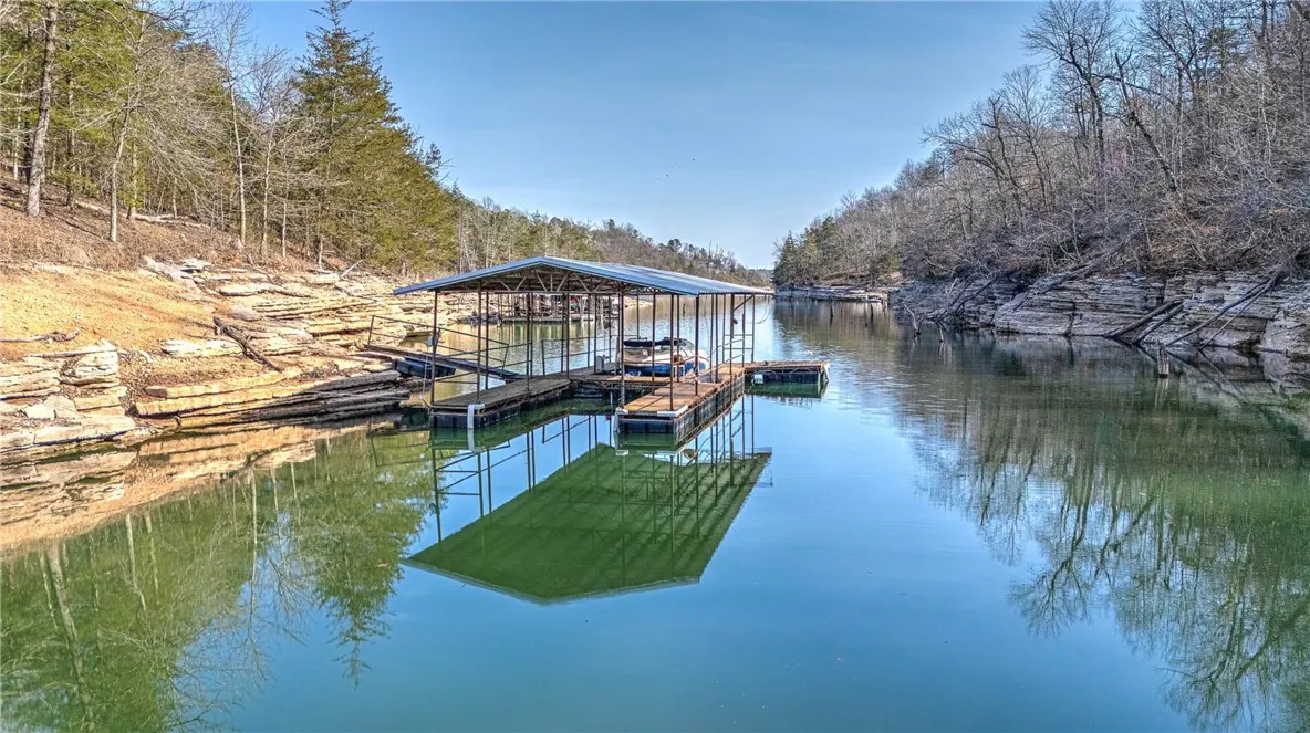 Scenic lake view with a covered boat dock, calm water reflecting the structure, and tree-lined shores under a clear blue sky.