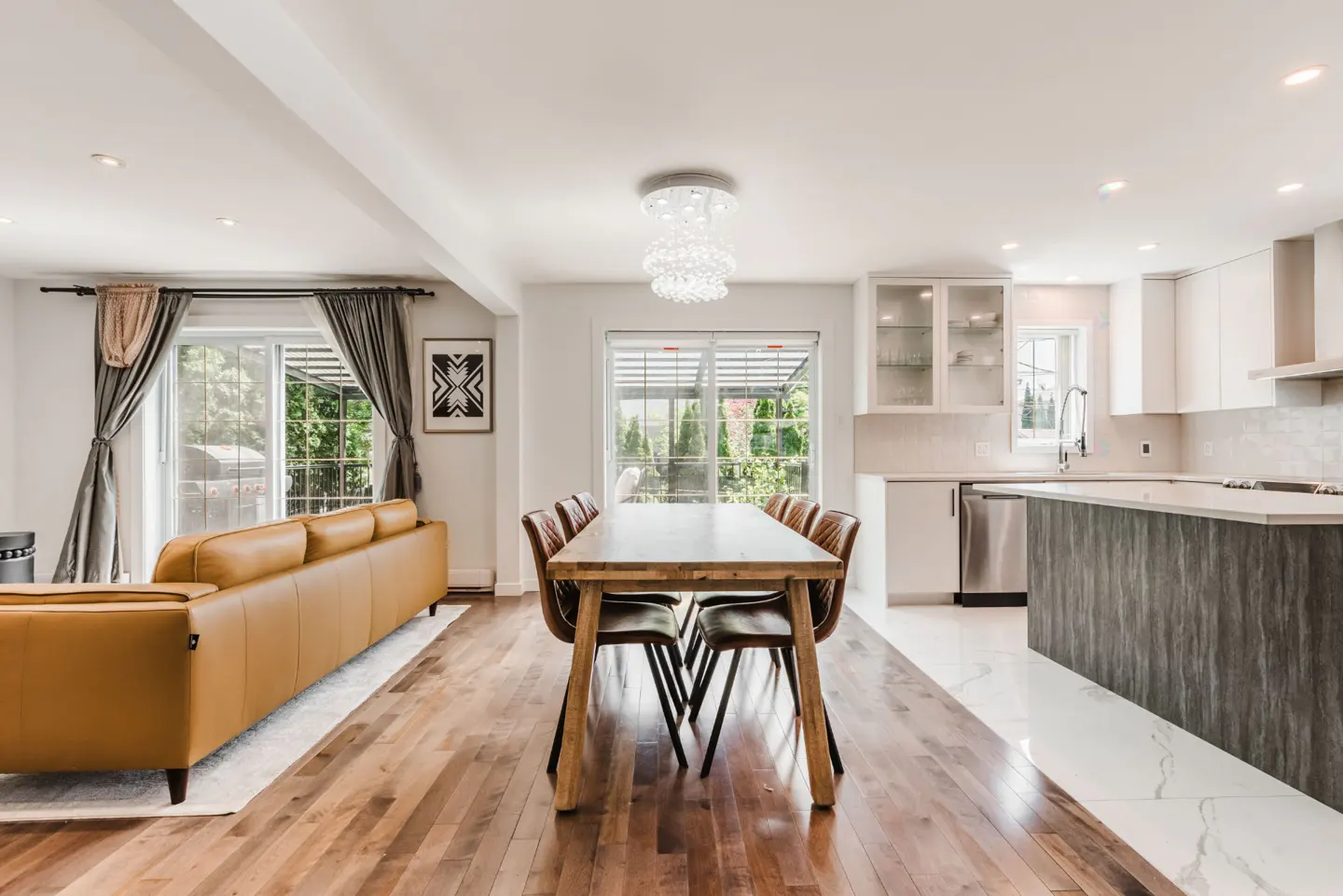 Open-concept living area with a tan leather sofa, wooden dining table, and modern kitchen with white cabinets and island.