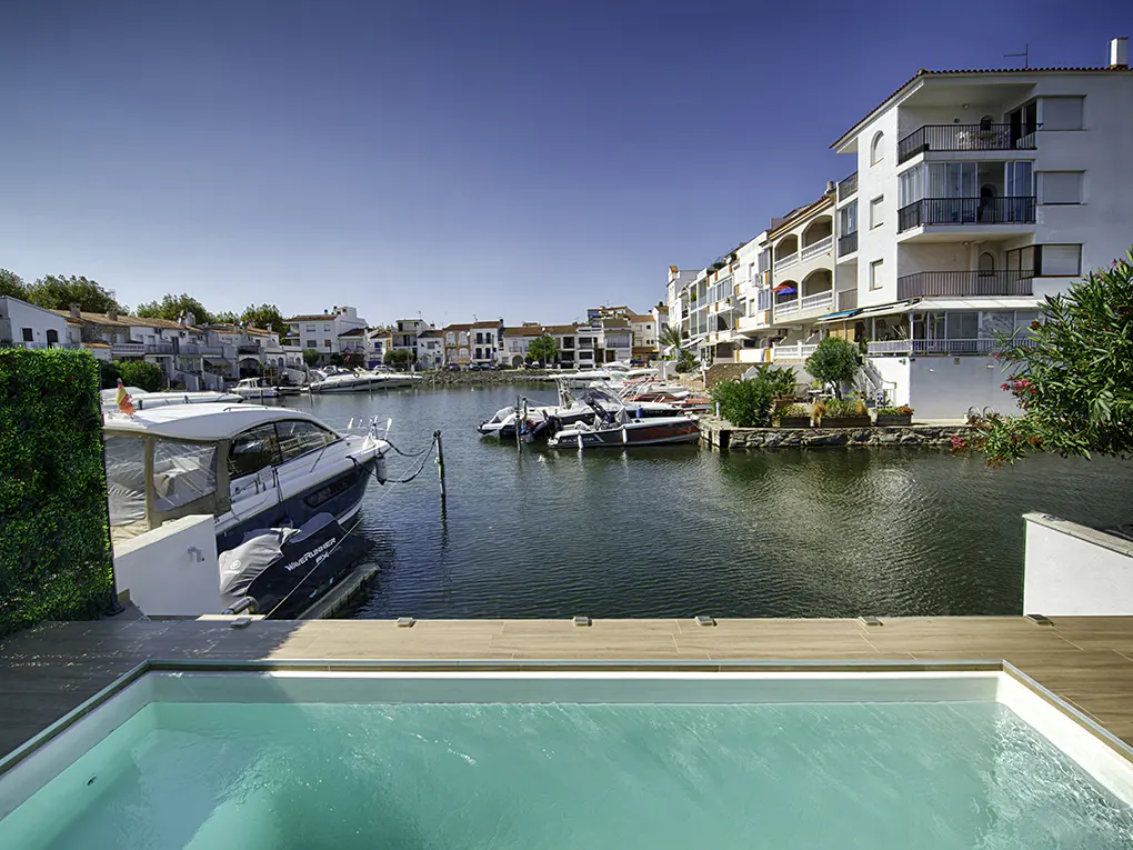 View from a pool overlooking a canal with boats docked in front of white buildings under a clear blue sky.
