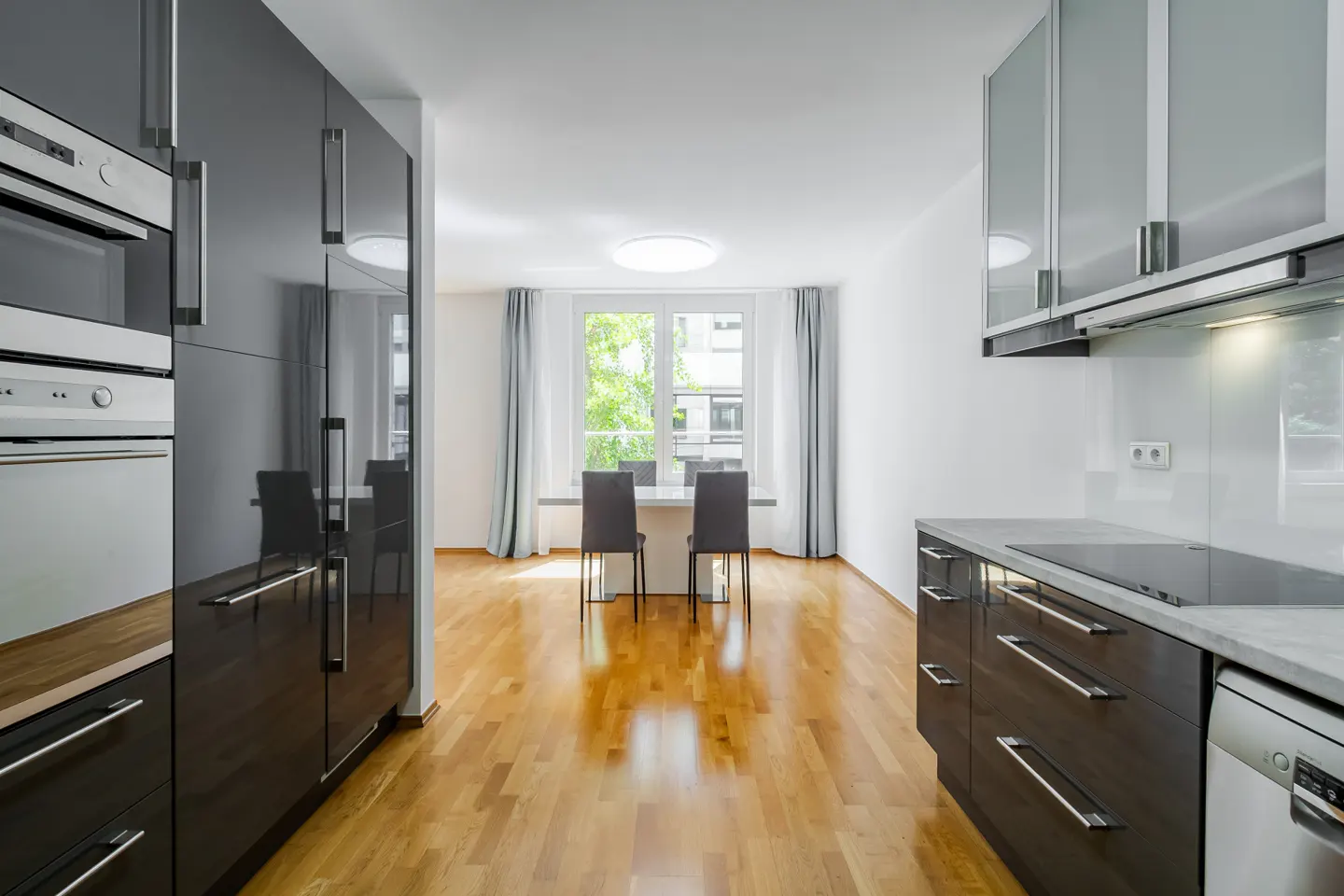 A modern kitchen with dark cabinets, stainless steel appliances, and a dining area with a table and chairs. Hardwood floors and a window with curtains.