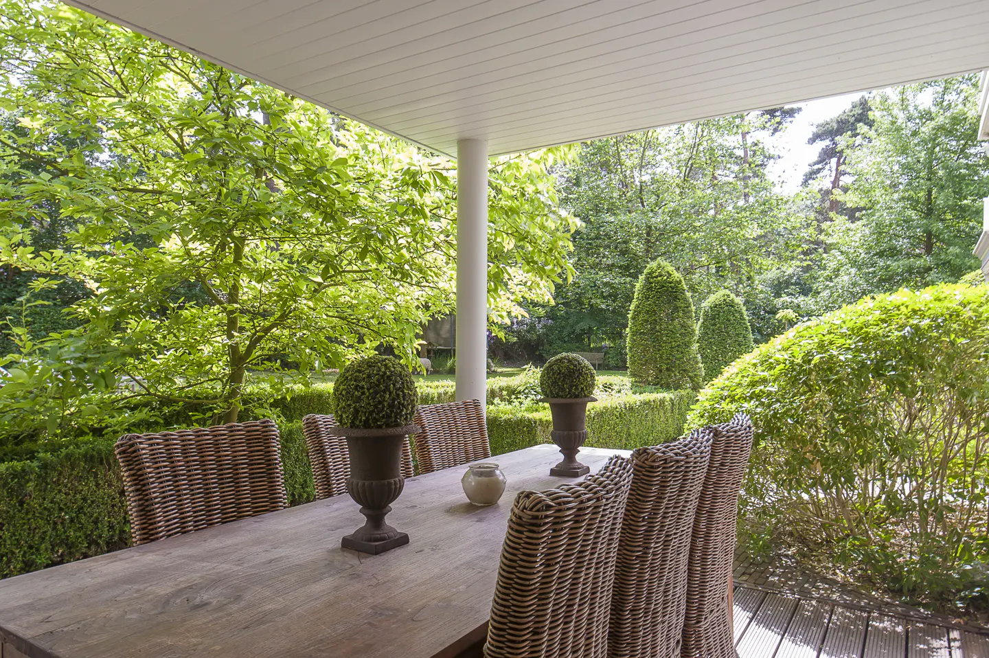 Covered patio with a wooden table, wicker chairs, and green garden view.