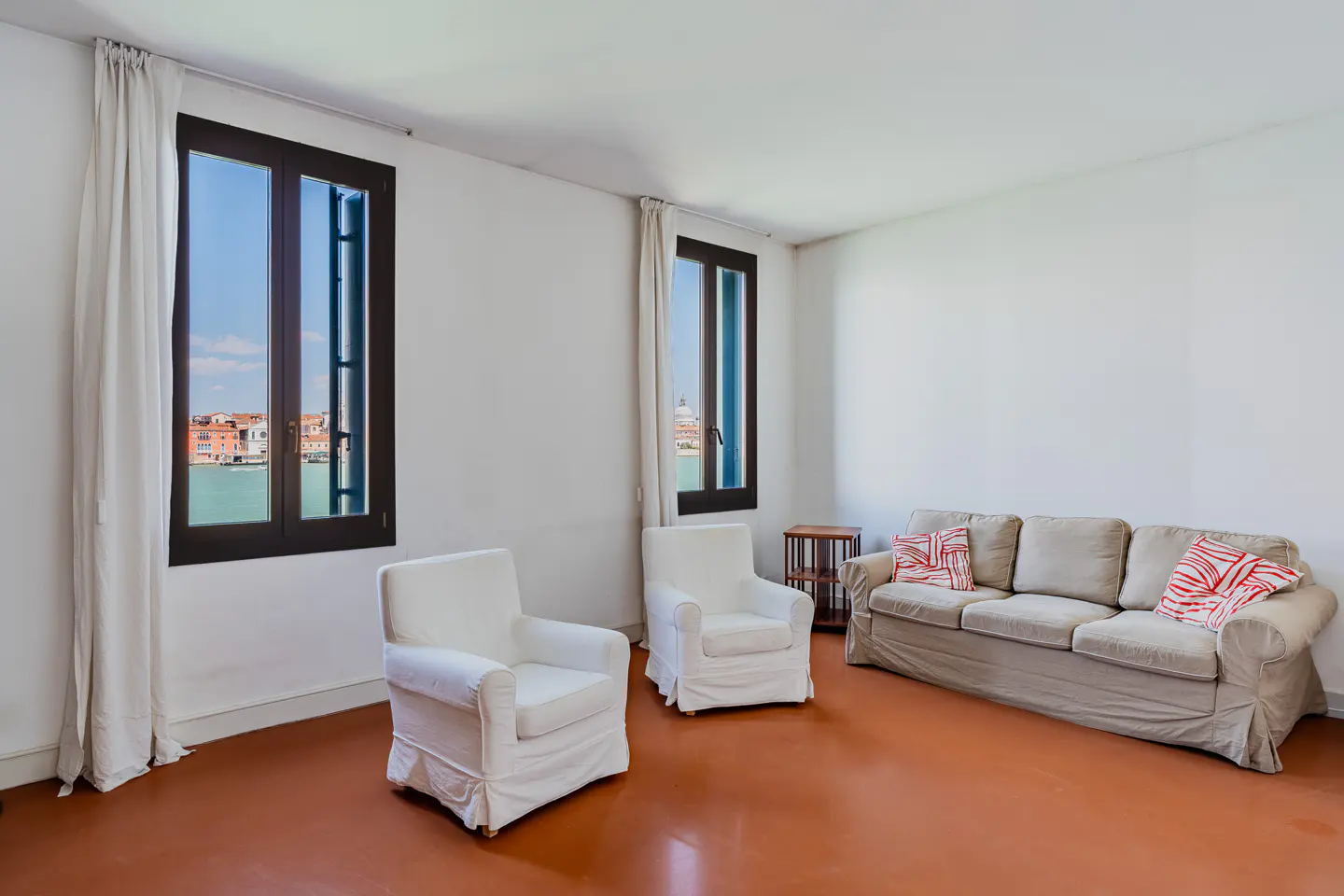 Bright room with two white armchairs, a beige sofa with red pillows, and two windows overlooking a canal in Venice.