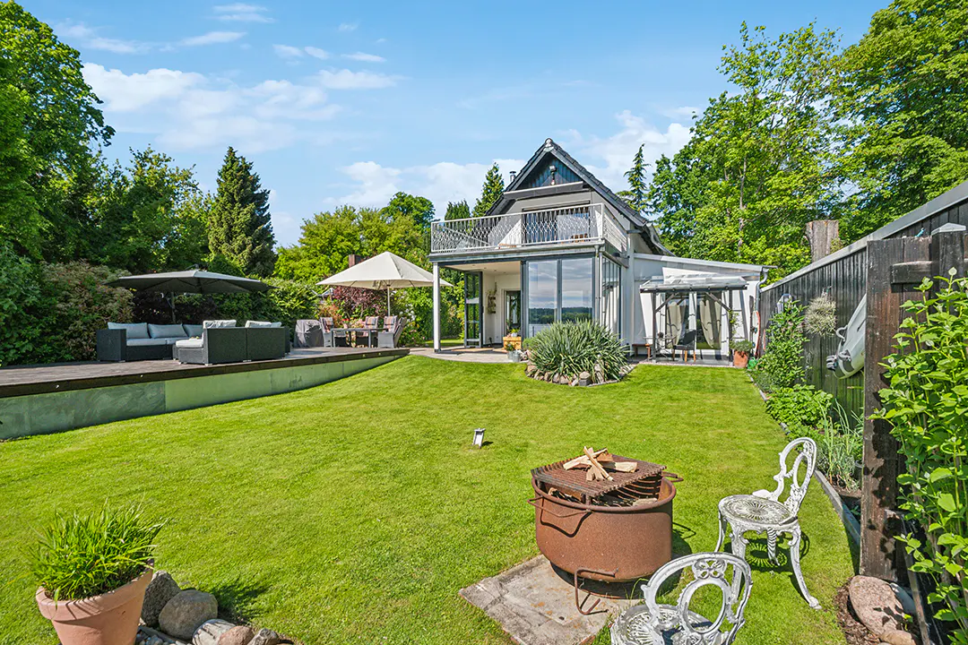 A modern house with a balcony overlooks a green lawn with a fire pit and patio furniture.