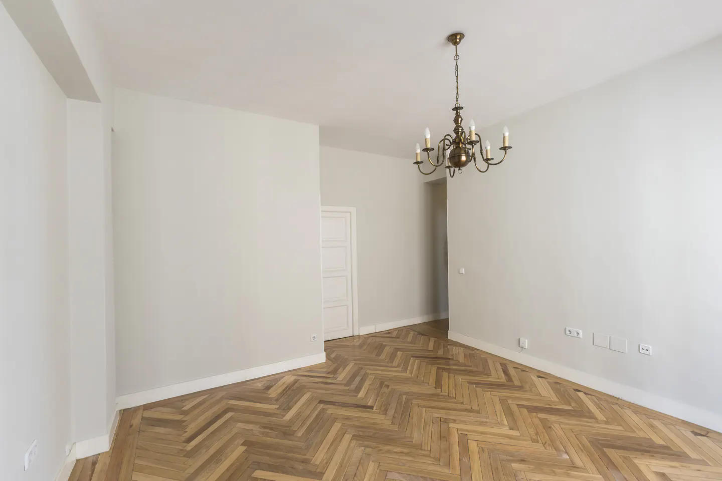 Empty room with light gray walls, herringbone wood floors, and a classic brass chandelier hanging from the ceiling.