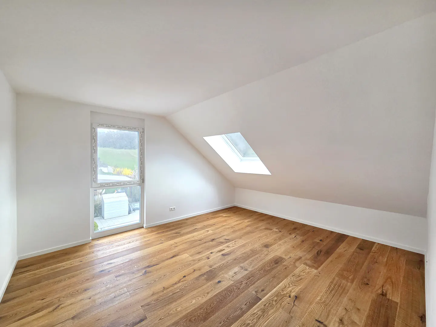 Bright attic room with white walls, wood floors, and a skylight. A window shows a green outdoor view.