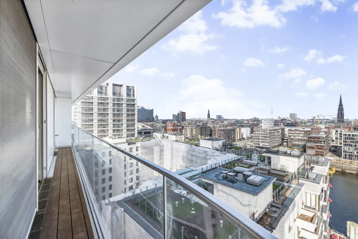 View from a modern balcony with glass railings overlooking a cityscape under a blue sky with scattered clouds.