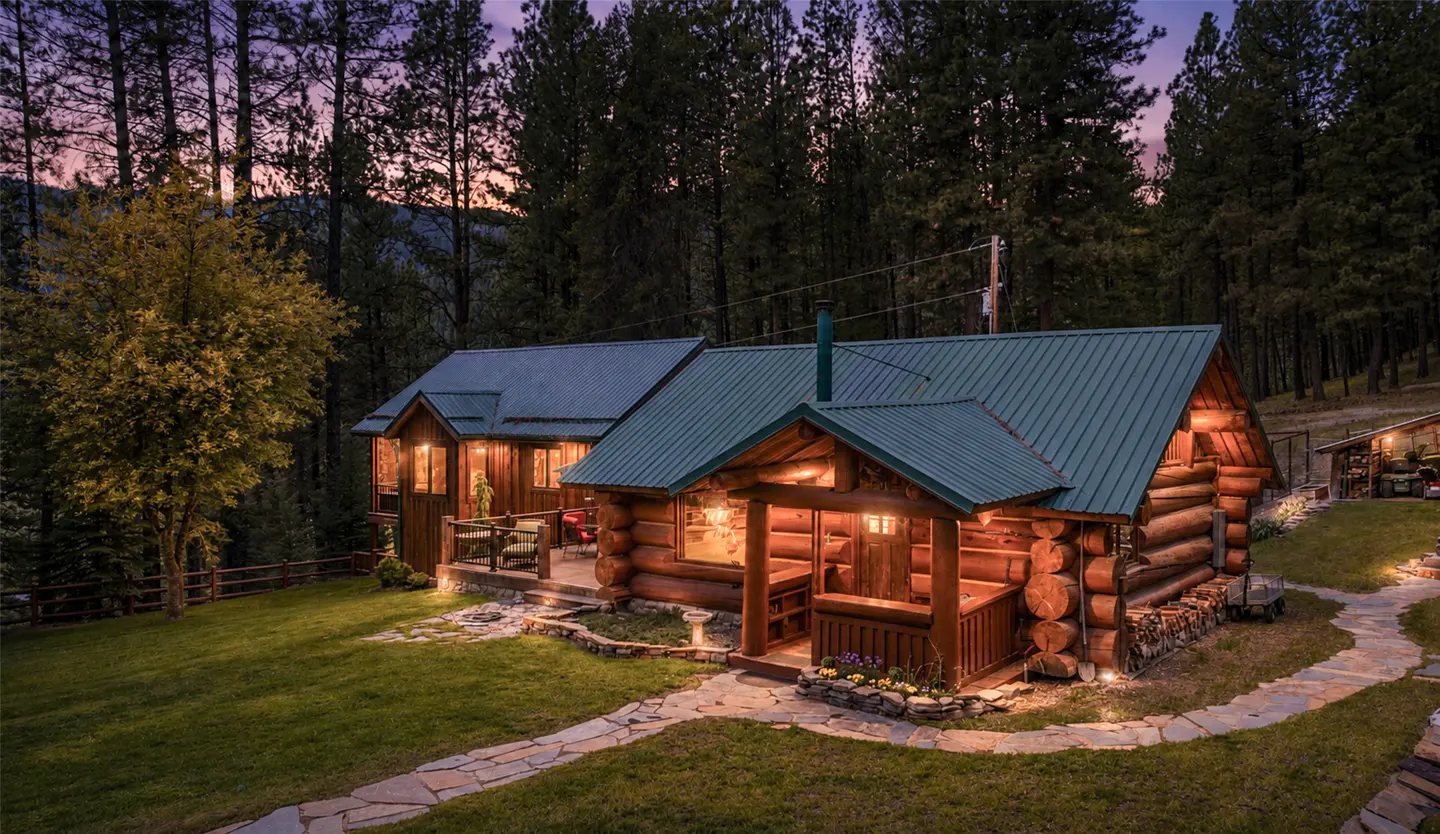 A log cabin with a green metal roof is nestled in a forest at dusk. A stone path leads to the porch, illuminated by warm lights.