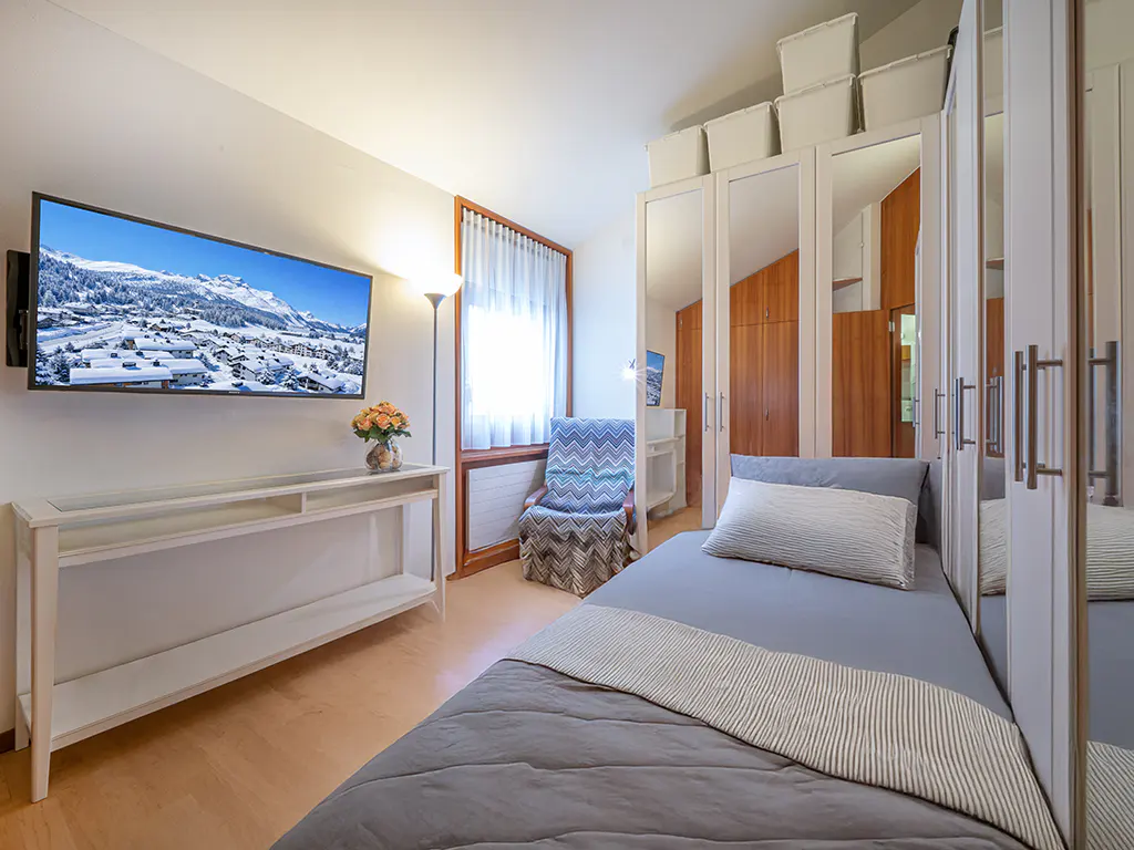 Bedroom with a gray bed, white console table, and a TV showing a snowy mountain scene. Mirrored closet doors and a patterned chair are also visible.
