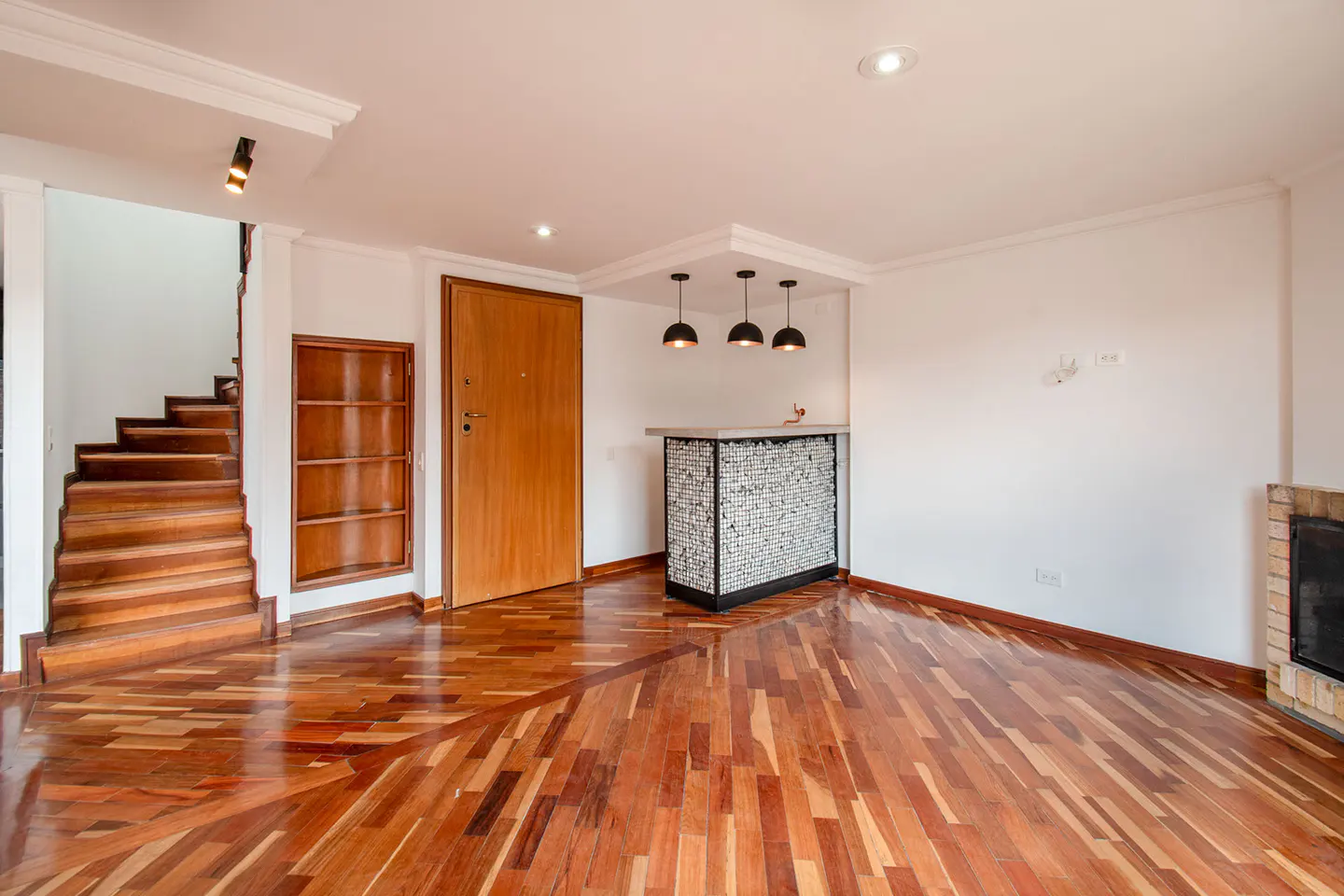 Modern living area with hardwood floors, a wooden staircase, a stone-fronted bar with pendant lights, and a brick fireplace.