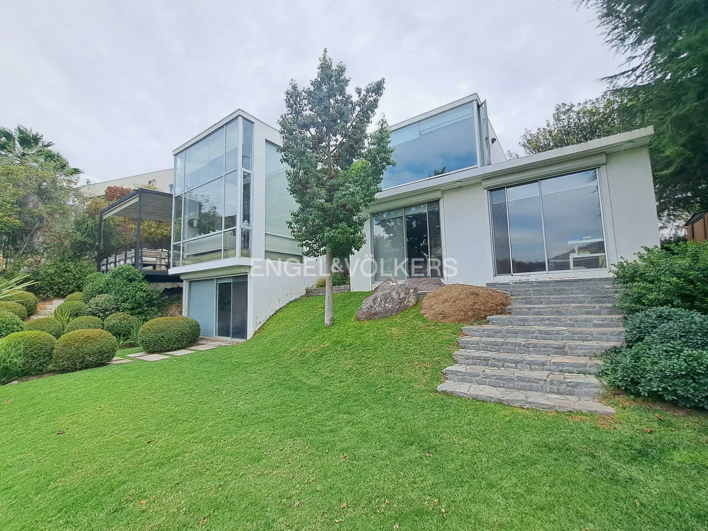Modern white house with large windows, green lawn, and stone steps leading up to the entrance. Engel & Volkers logo visible.