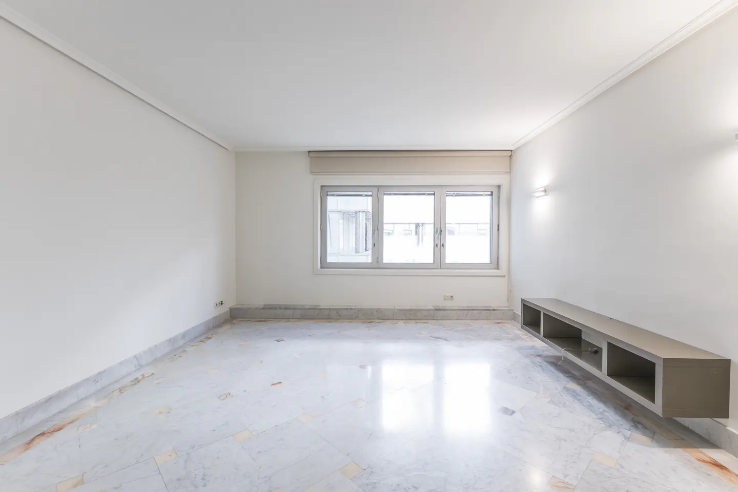 Bright, empty room with white walls, marble floor, and a window. A long, gray shelf is mounted on the right wall.