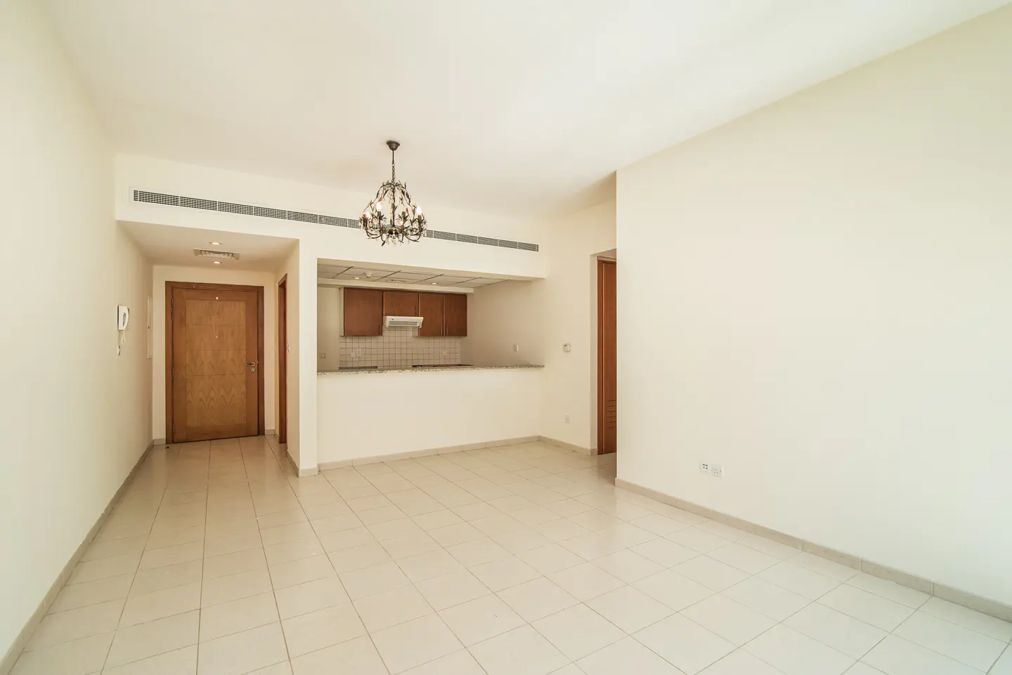 An empty apartment with cream walls, tile floors, and a chandelier. The kitchen has brown cabinets and a white countertop.