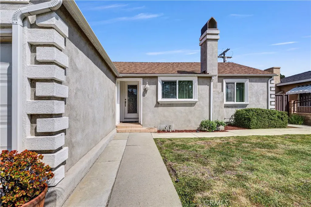 Exterior view of a one-story stucco house with a brown roof, white trim, and a chimney under a blue sky.