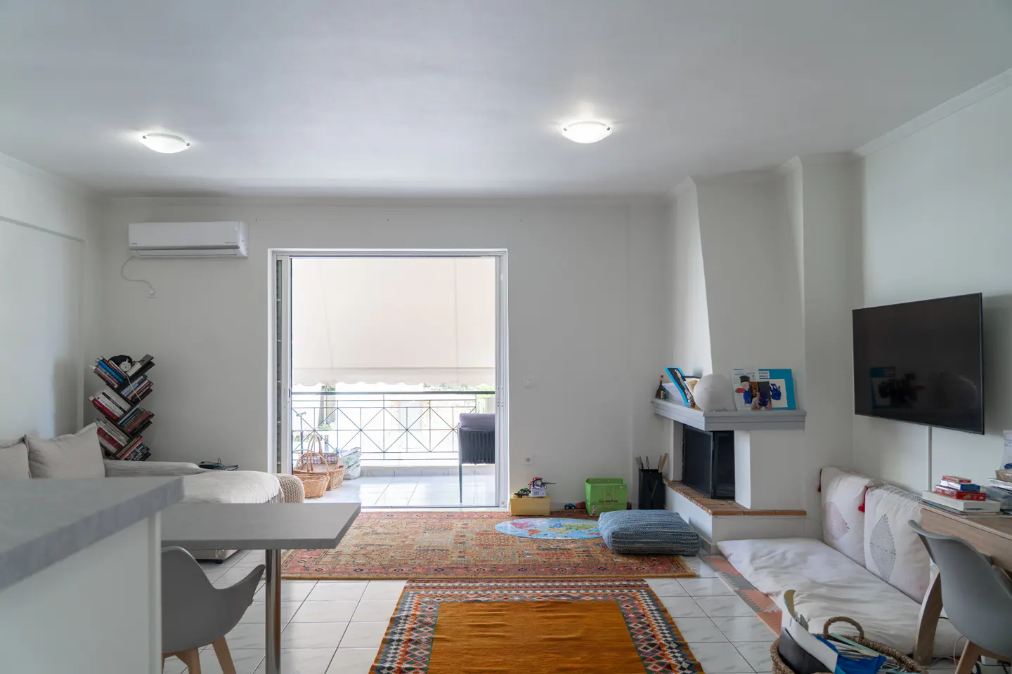 Bright living room with white walls, tiled floors, patterned rugs, a fireplace, wall-mounted TV, and balcony access.