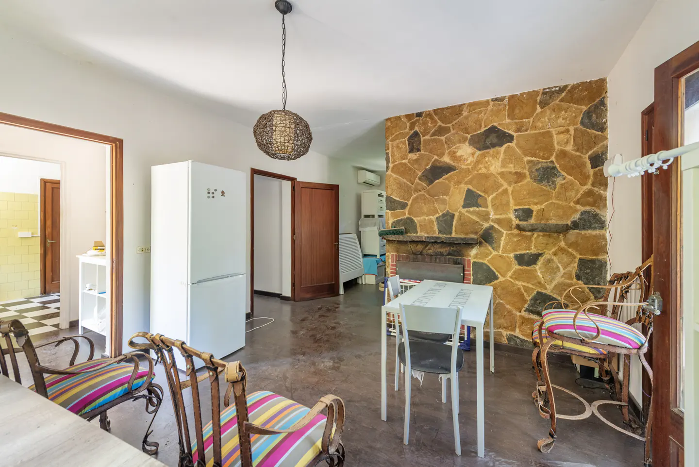 Dining area with a stone wall, white refrigerator, dining table with chairs, and colorful striped cushions.