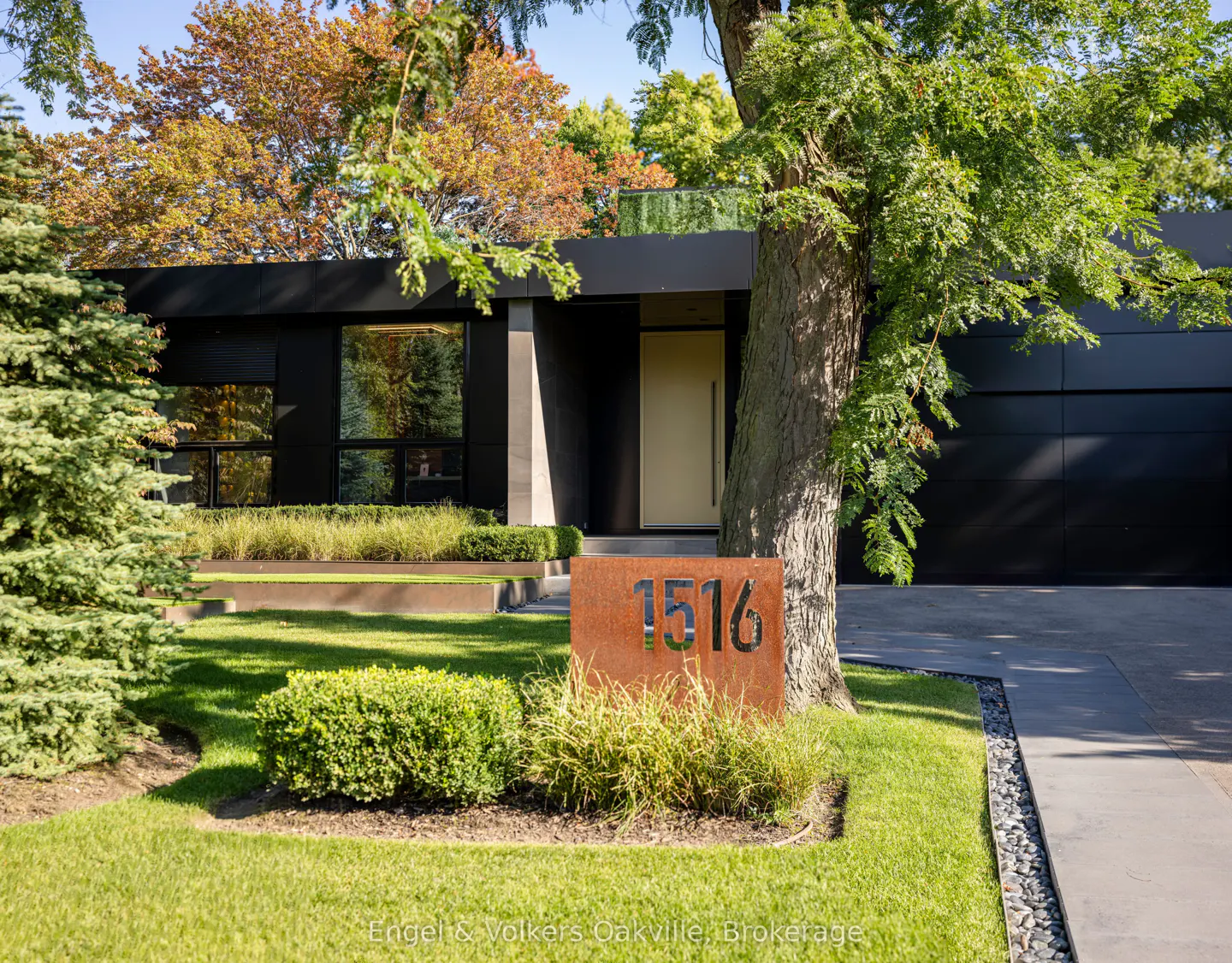 Modern black house with a beige door, green lawn, and a rusty metal address sign reading "1516". A large tree stands to the right.