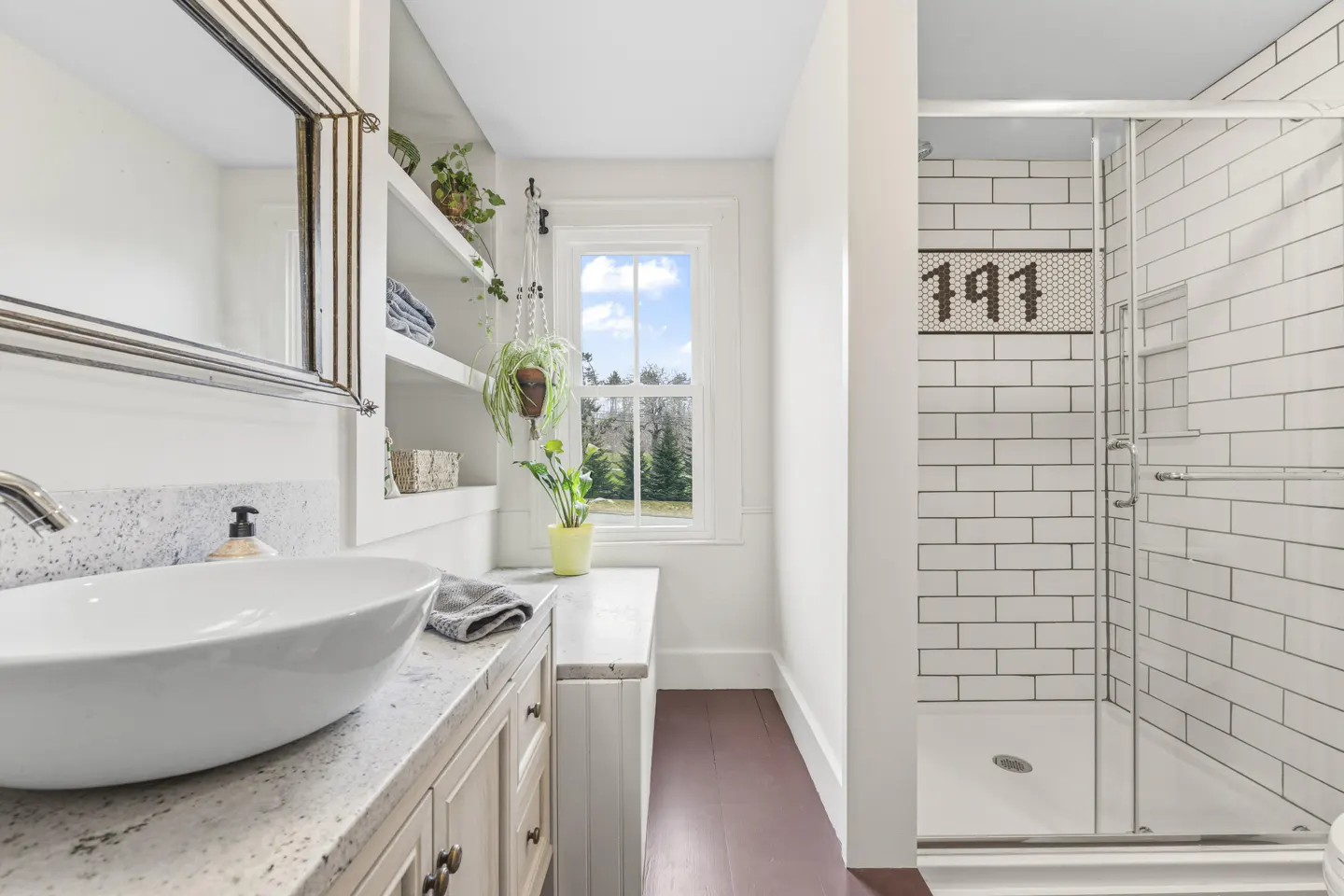 Bright bathroom with white walls, a white sink, and a glass shower with white subway tile and a black mosaic number "141".