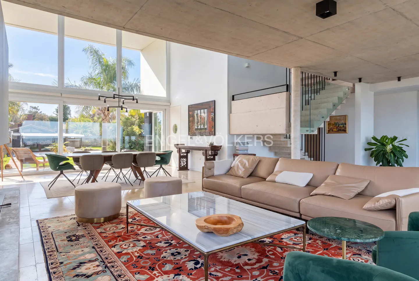Modern living room with a beige sofa, marble coffee table, red patterned rug, and dining area with floor-to-ceiling windows.