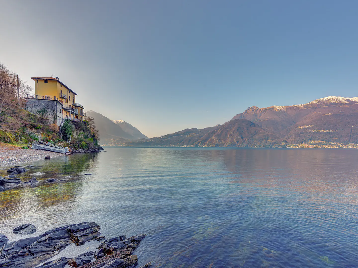 Lake Como view with a yellow house on a hill, a rocky shore, and mountains in the background under a clear blue sky.